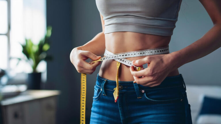 Uma mulher com uma fita métrica medindo a cintura, com uma calça jeans e um top cinza, mostrando que suplemento termogênico emagrece.