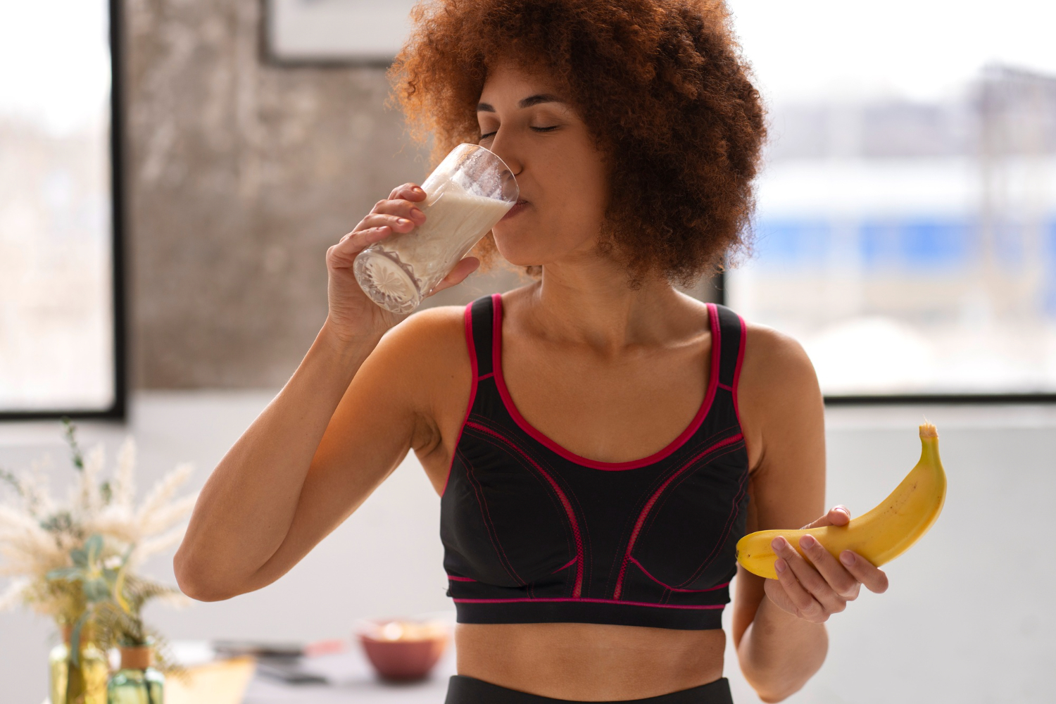 Uma mulher com roupa de ginástica tomando uma vitamina de creatina com whey protein em uma mão, e na outra, segurando uma banana dentro de uma sala.