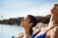 duas mulheres de biquíni deitadas em uma espreguiçadeira tomando sol em frente ao mar com um bronzeamento saudável de betacaroteno