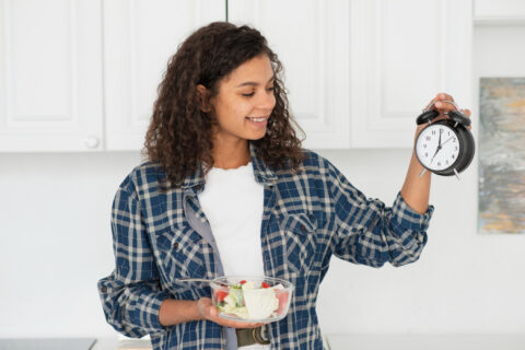 Uma menina na cozinha segurando um relógio em uma mão e na outra mão uma vasilha com salada, simulando o tempo do jejum intermitente