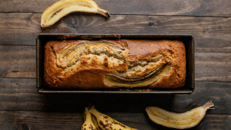 Bolo de whey com banana em cima de uma bancada de madeira com pedaços de banana in natura em volta