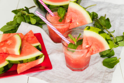 Dois drinks sem álcool decorados com fatias de melancia e folhas de hortelã ao lado de um prato com pedaços de melancia em uma mesa branca.