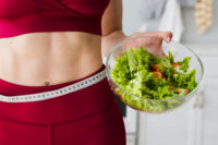 Uma mulher de roupa de ginástica com uma fita métrica na cintura e um pote de salada na mão, simulando estratégias de redução de percentual de gordura