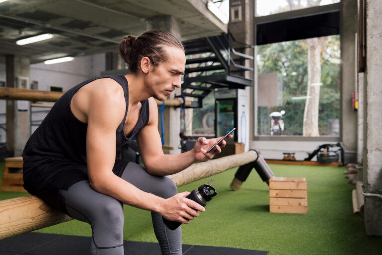 Homem sentado com roupa de ginástica na academia olhando para o celular e vendo sobre como montar um treino eficiente enquanto segura uma garrafa de água na outra mão