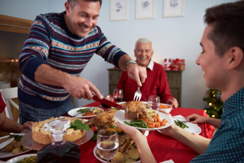 Família reunida no Natal, com um homem feliz servindo peru e vegetais em um ambiente decorado para as festas de fim de ano saudáveis.