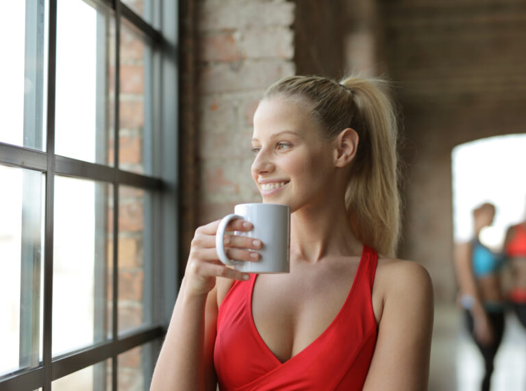 uma mulher loira sorrindo com um top vermelho de academia segurando uma xícara de café com creatina