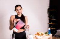 Mulher jovem vestindo roupas esportivas pretas, segurando um liquidificador com um shake de whey protein rosa enquanto despeja em um copo.