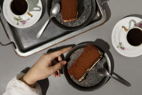 Mão segura prato de tiramisù proteico ao lado de xícara de café, em comemoração ao dia mundial do café.