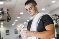 Jovem na academia com uma toalha sobre os ombros, preparando um shake com suplemento, enquanto se questiona se o melhor suplemento é o hipercalórico ou whey.