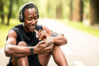 homem negro comendo barrinha de proteína sentado num parque olhando o celular e ouvindo música